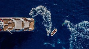 Aerial view of the luxury superyacht JoyMe at anchor with a tender boat and a jet ski creating wakes in the deep blue ocean, surrounded by sparkling water.